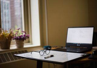 A computer on a desk with a whiteboard laying on its side in front of it and glasses on top of the whiteboard.